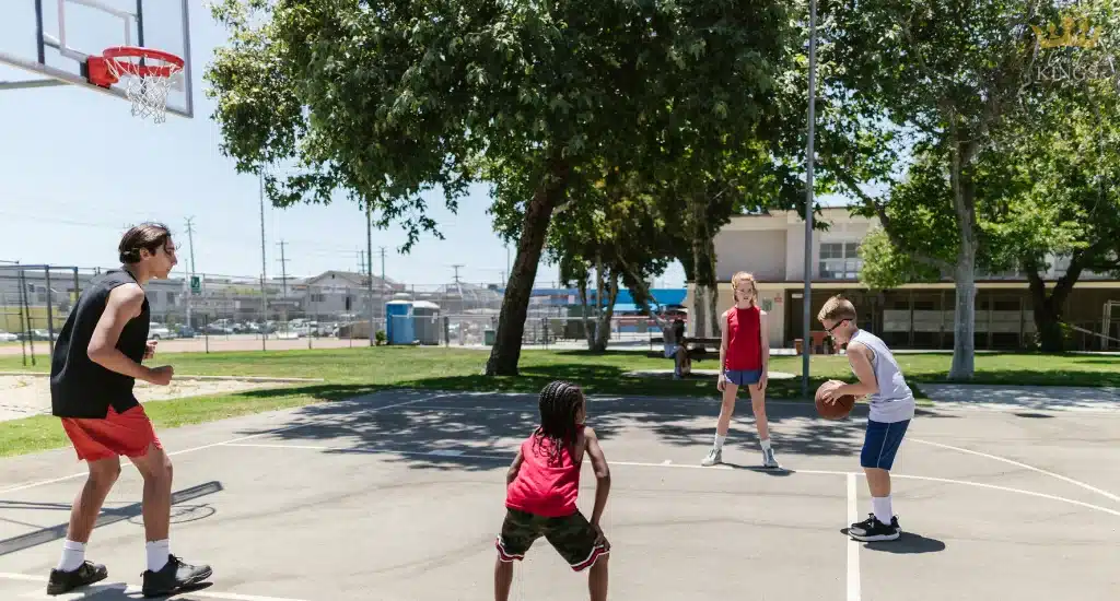 Kids playing basketball on sunny outdoor court near school.