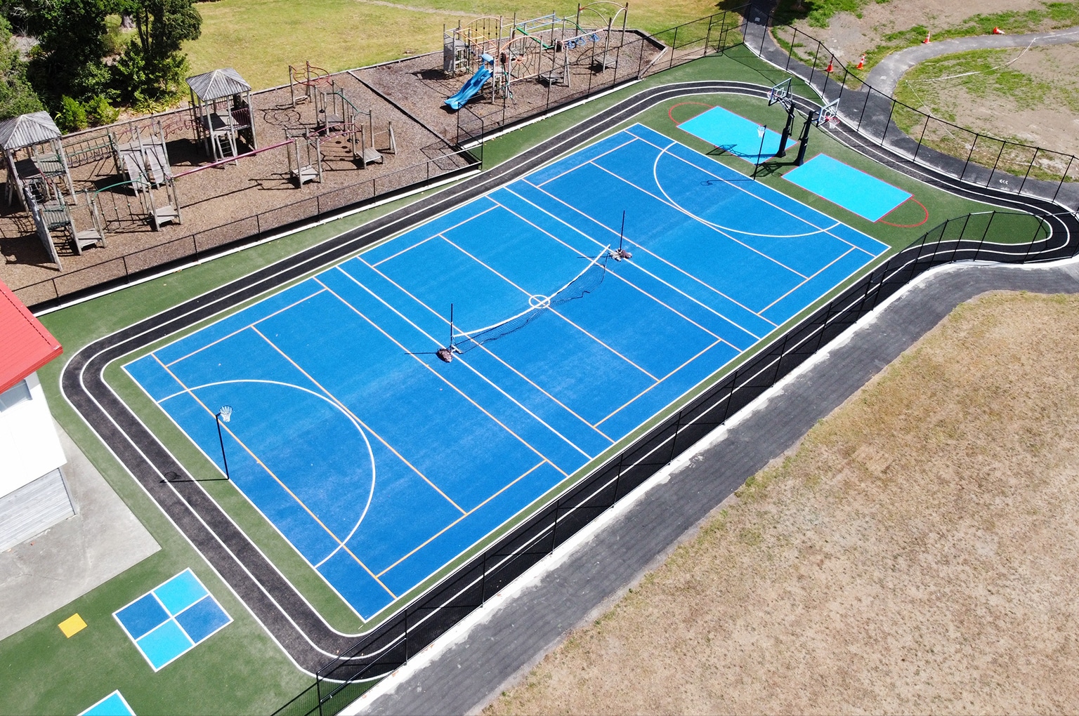 Aerial view of a blue multi-sport court with basketball hoops, netball posts, and a nearby playground.