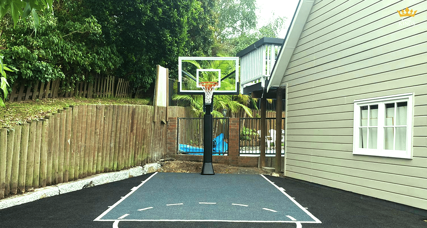 Outdoor basketball hoop on a compact driveway court beside a house, with timber fence and trees in the background