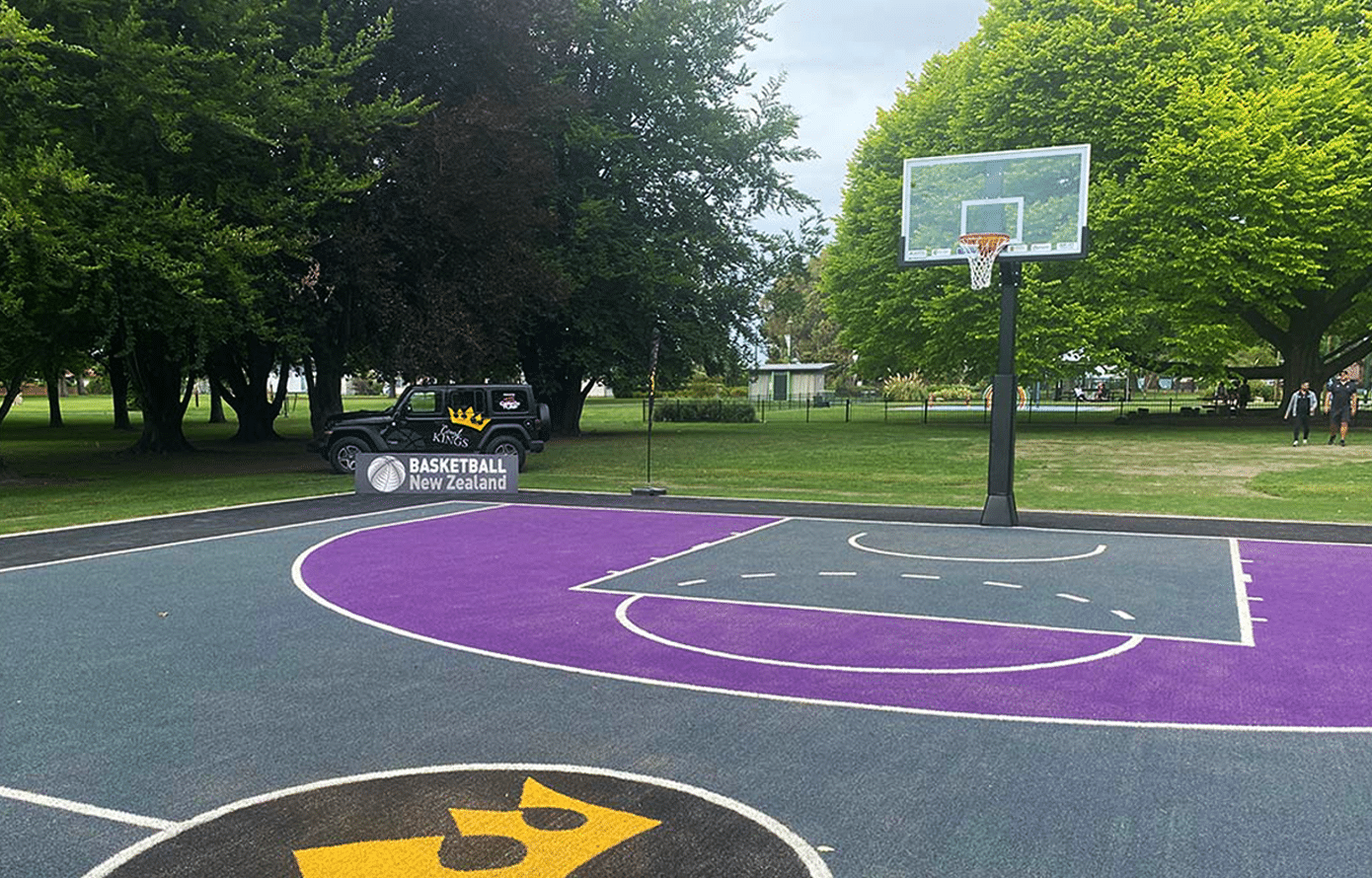 A professional in-ground basketball hoop with a clear glass backboard, installed by Court Kings on a custom purple and grey outdoor court in a New Zealand park.